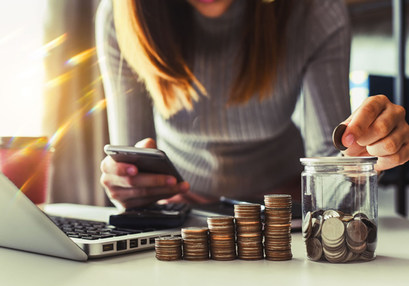 Computador e calculadora sobre a mesa, mulher segurando celular colocando moedas em um pote, ao lado pilhas crescentes de moedas